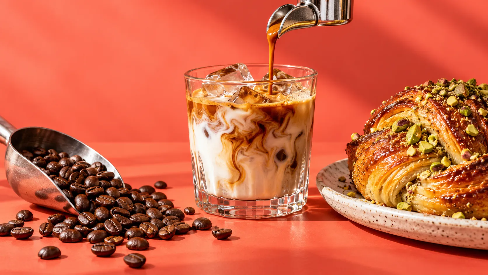 Espresso pouring into an iced latte beside coffee beans and a pistachio bun on a coral table